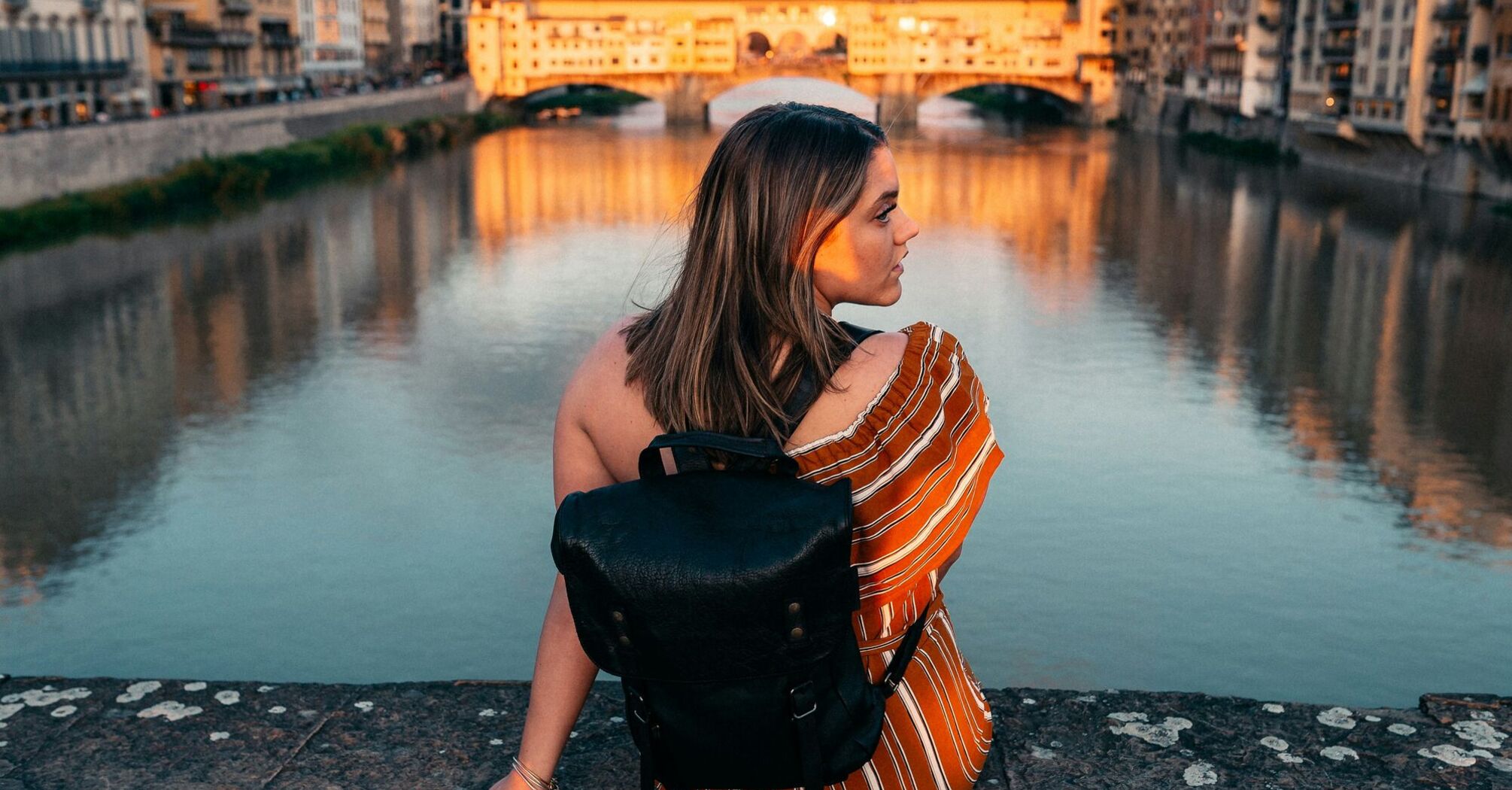 woman in white and orange striped shirt and black leather sling bag siting on gray concrete