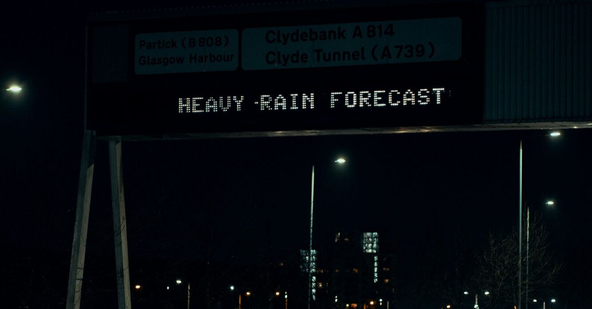 A digital road sign at night warning of heavy rain, with wet roads and passing cars beneath