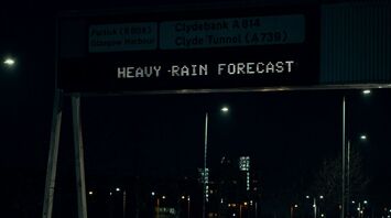 A digital road sign at night warning of heavy rain, with wet roads and passing cars beneath