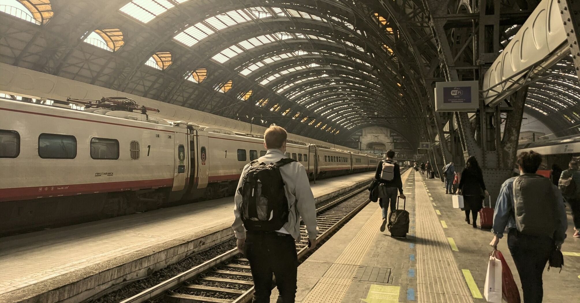 A group of travelers walking along a platform in a Milano Centrale train station with a train waiting at the platform