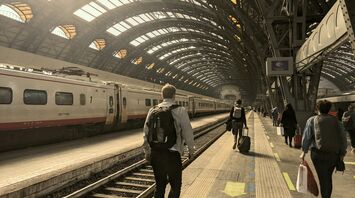 A group of travelers walking along a platform in a Milano Centrale train station with a train waiting at the platform