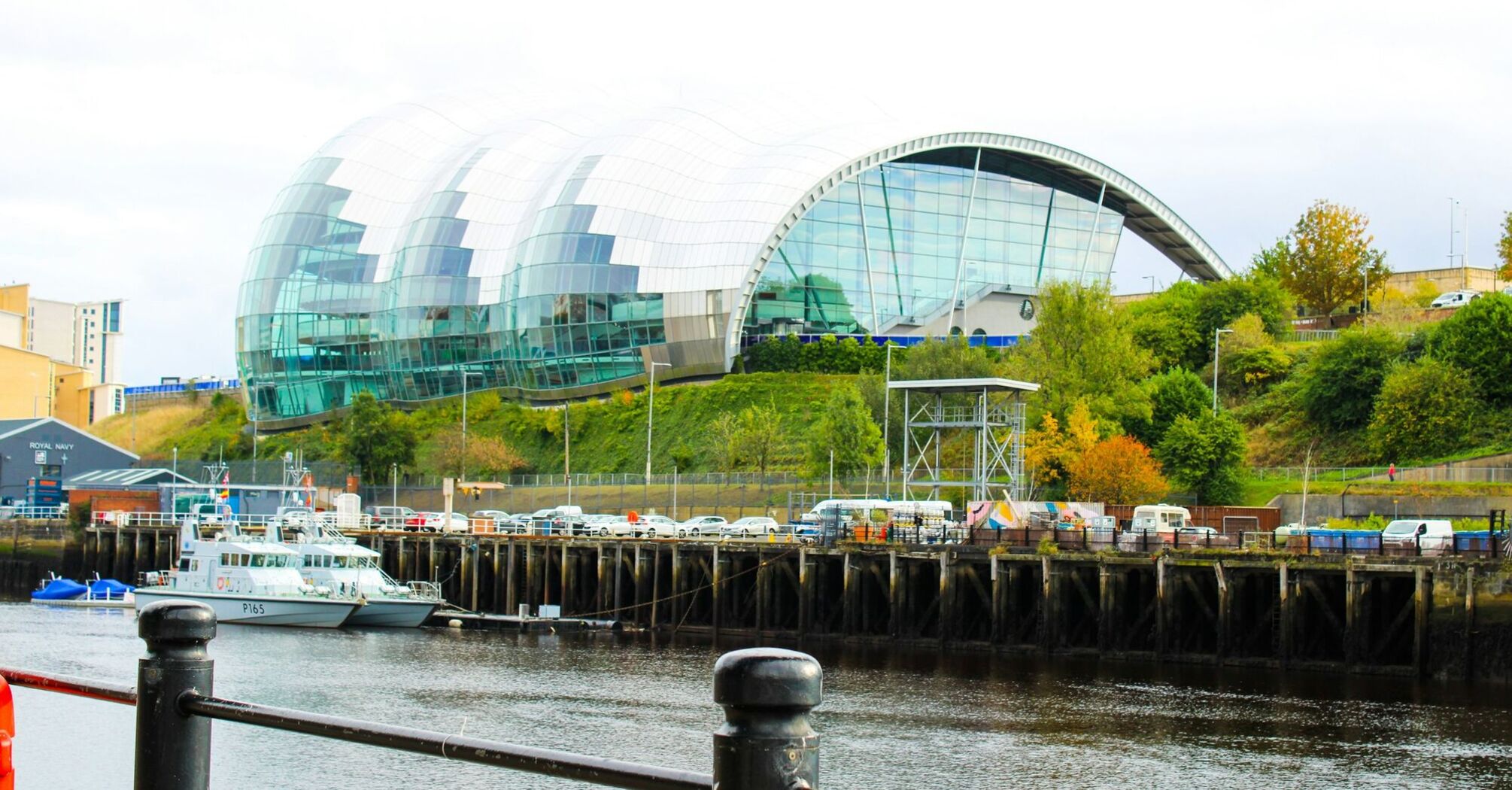 A modern, glass-covered building with a curved design overlooking a riverside in Gateshead, with boats docked along the water and greenery in the background