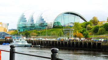 A modern, glass-covered building with a curved design overlooking a riverside in Gateshead, with boats docked along the water and greenery in the background