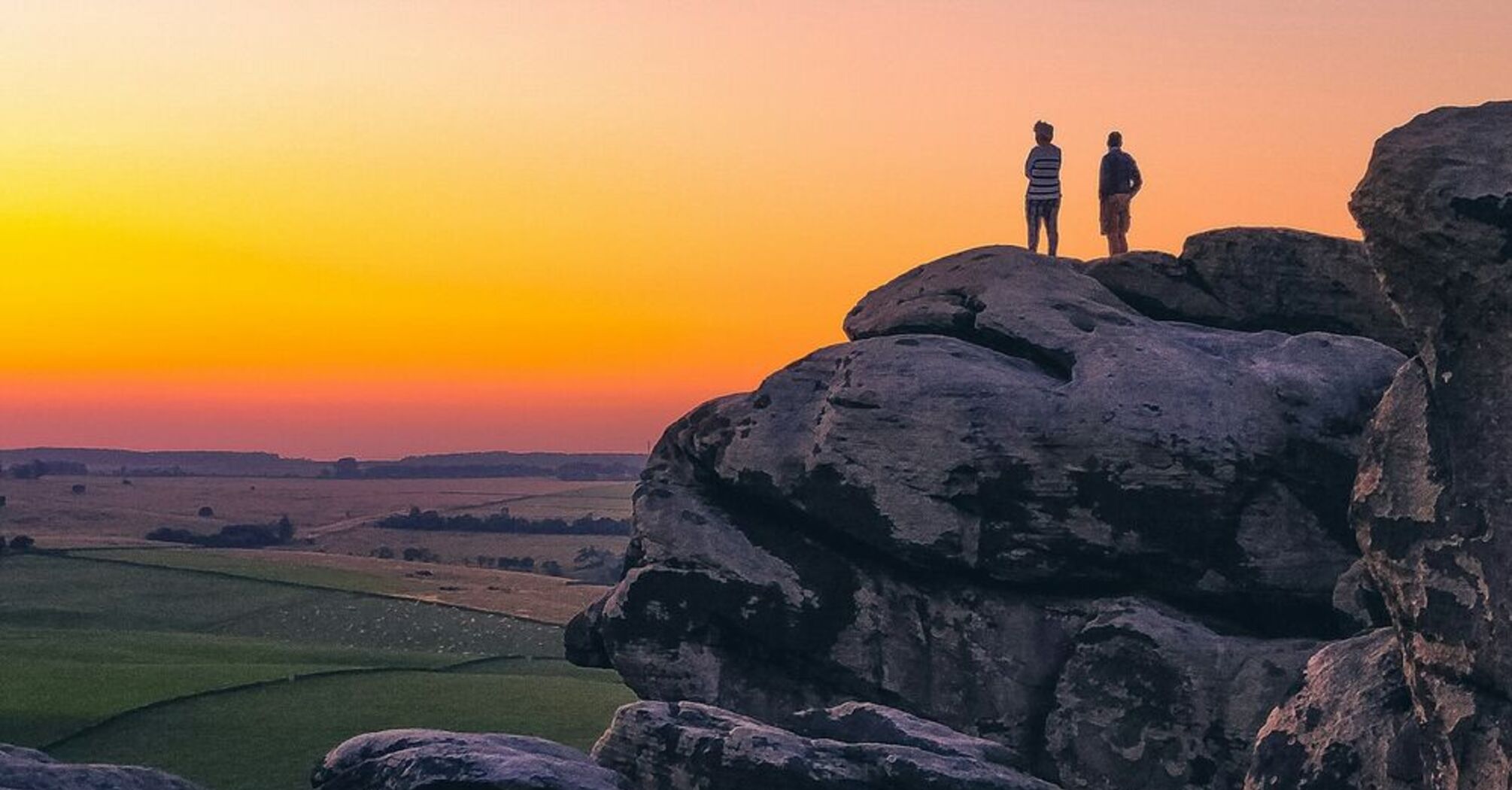 Two people standing on large rock formations at sunset, overlooking Yorkshire landscape with a gradient sky in the background