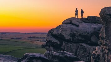 Two people standing on large rock formations at sunset, overlooking Yorkshire landscape with a gradient sky in the background