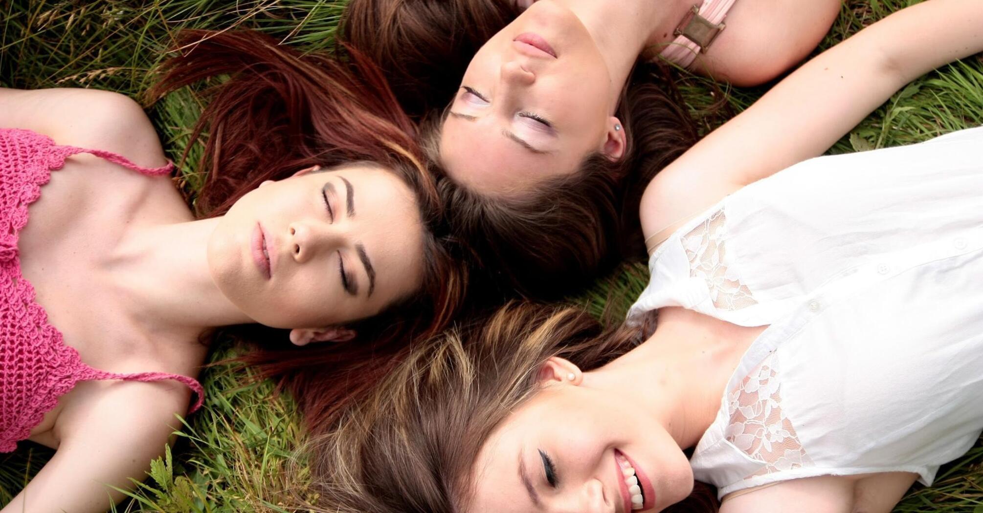 Three women lying on grass with their eyes closed, enjoying a peaceful moment outdoors