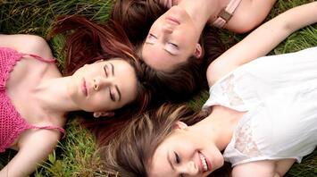 Three women lying on grass with their eyes closed, enjoying a peaceful moment outdoors