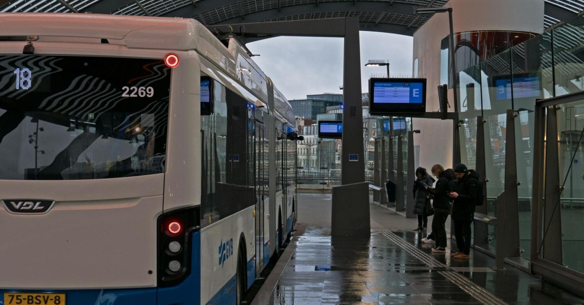 A bus at a modern station with a few passengers waiting on the platform, under a curved roof on a rainy day