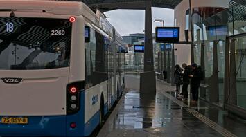 A bus at a modern station with a few passengers waiting on the platform, under a curved roof on a rainy day