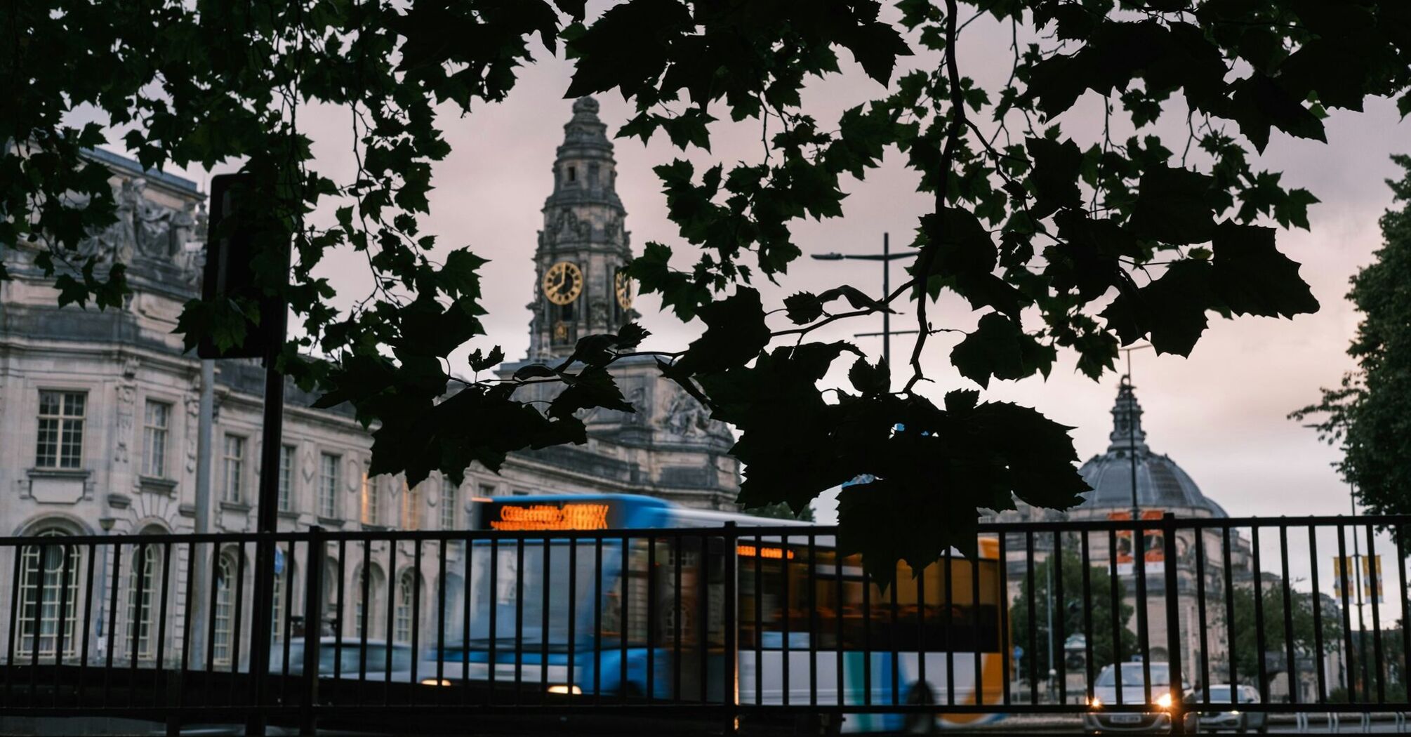 A Stagecoach bus passes by historic buildings in Cardiff, with a clock tower visible through the leaves of nearby trees