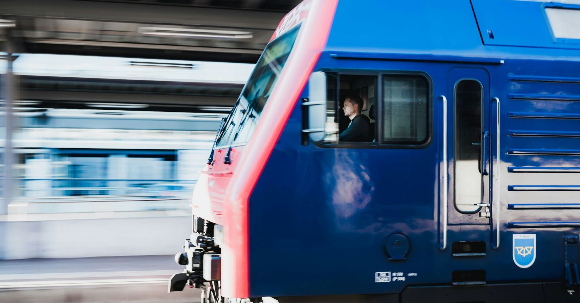 A blue and red train moving swiftly past a station with the driver visible in the cabin