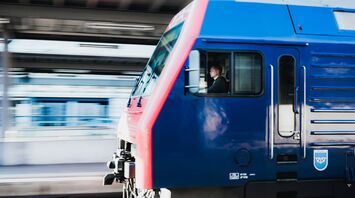 A blue and red train moving swiftly past a station with the driver visible in the cabin