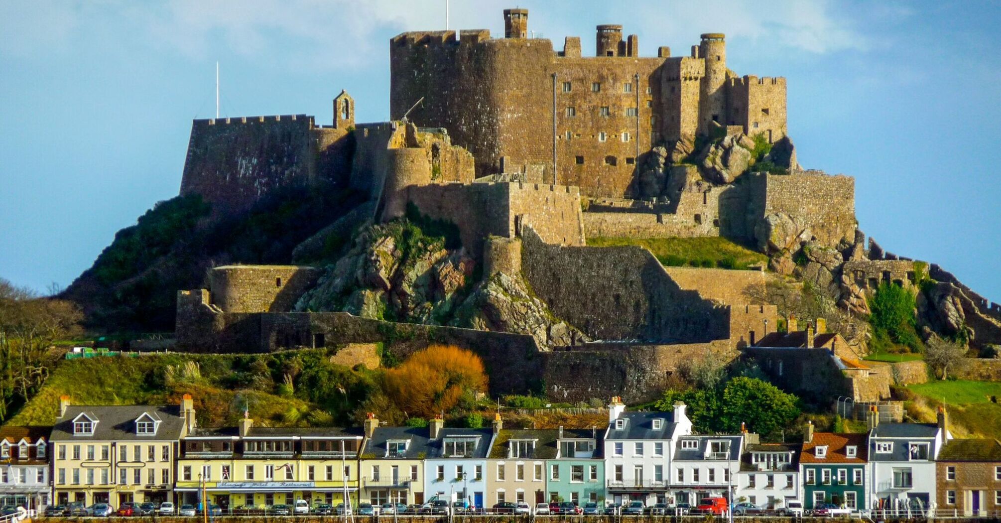 A historic castle in Jersey sits atop a hill overlooking a harbor with colorful houses and boats docked along the waterfront