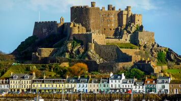 A historic castle in Jersey sits atop a hill overlooking a harbor with colorful houses and boats docked along the waterfront