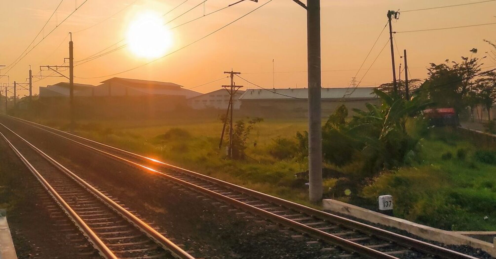 A railway track at sunrise with overhead wires, extending towards the horizon in a rural area