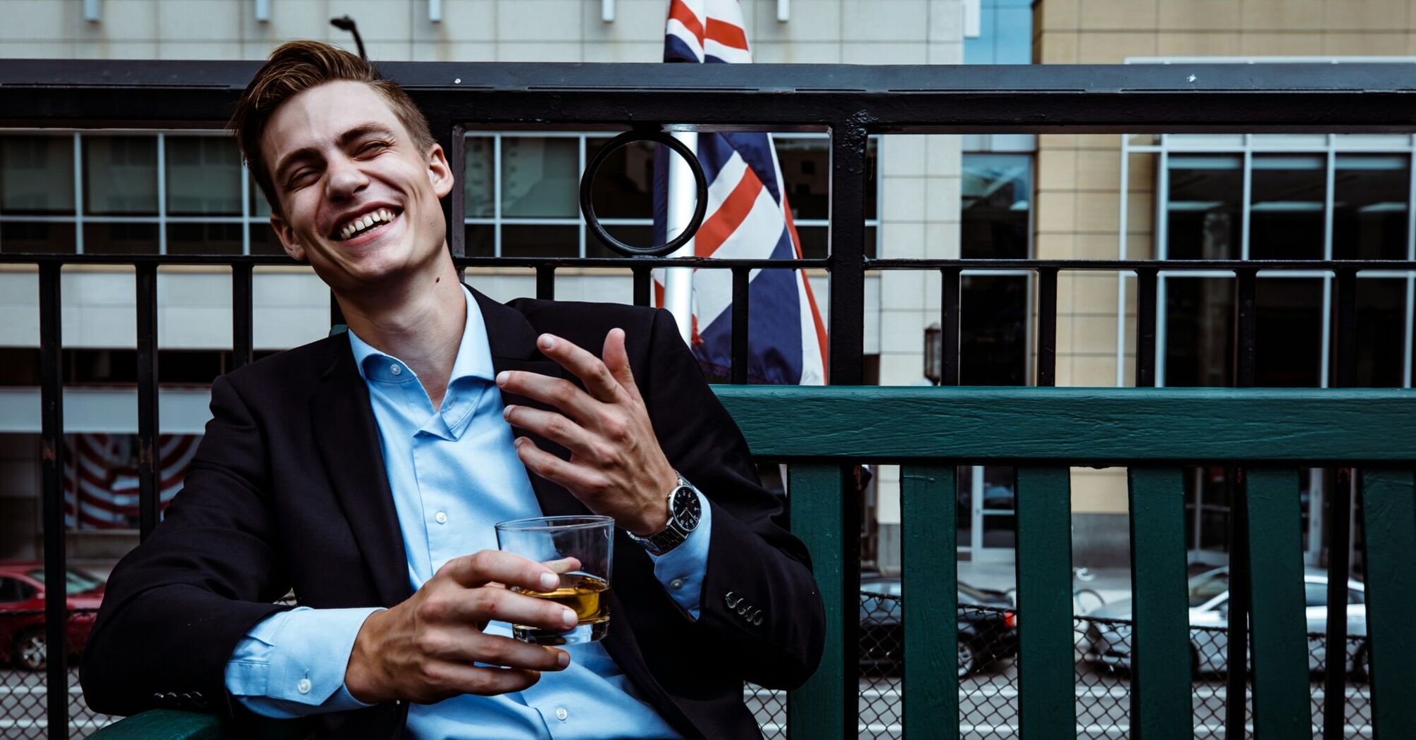 man smiling while sitting and holding whisky glass near concrete building