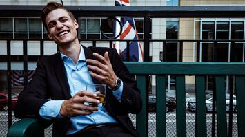 man smiling while sitting and holding whisky glass near concrete building