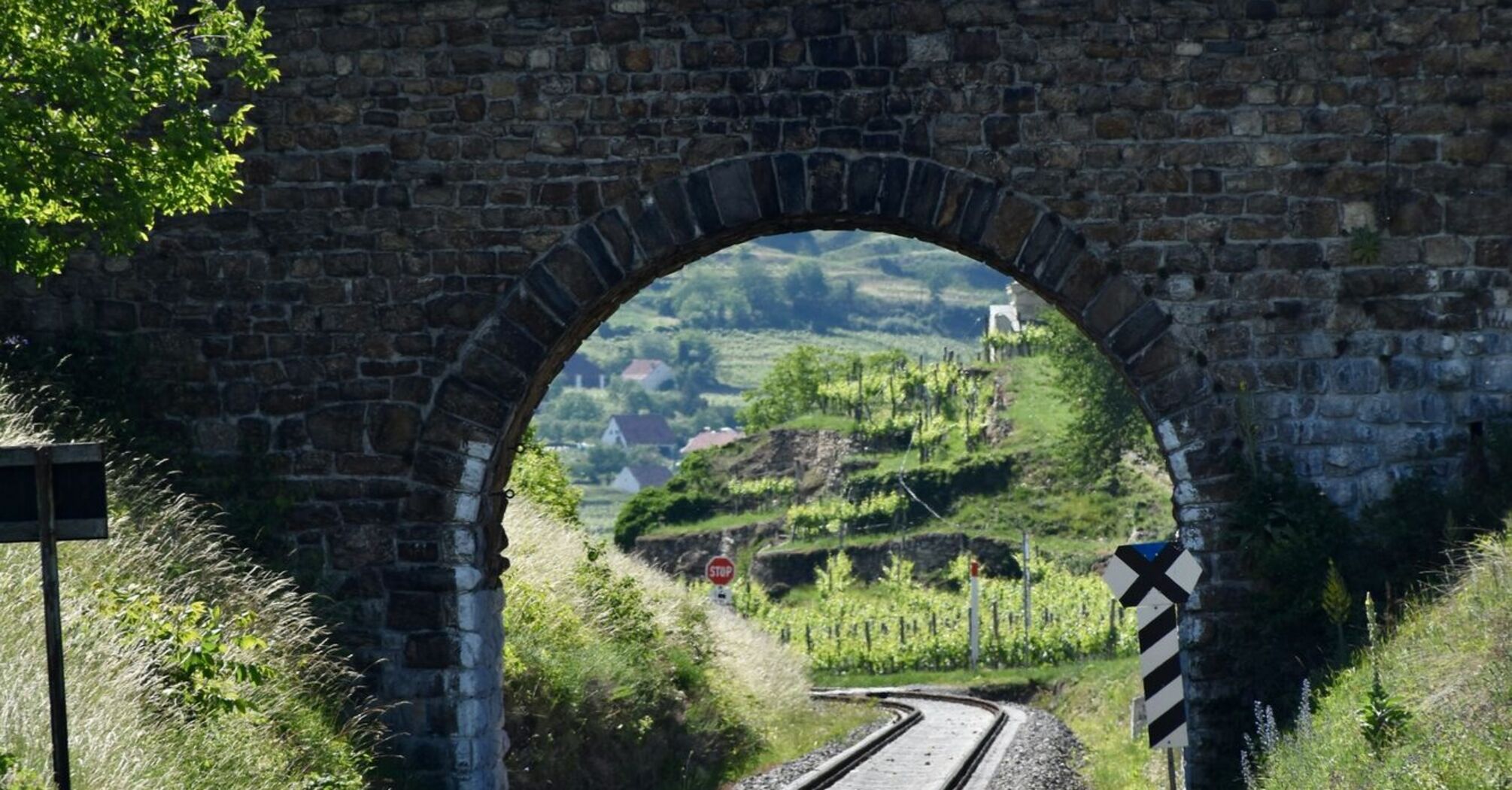 A stone railway bridge with an arch over train tracks, surrounded by lush green hills under a cloudy sky