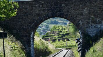 A stone railway bridge with an arch over train tracks, surrounded by lush green hills under a cloudy sky