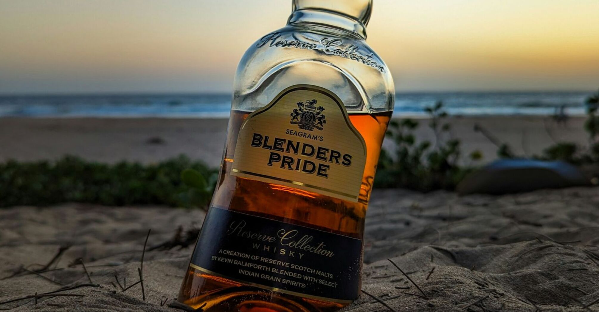 A bottle of whiskey partially buried in the sand at the beach during sunset