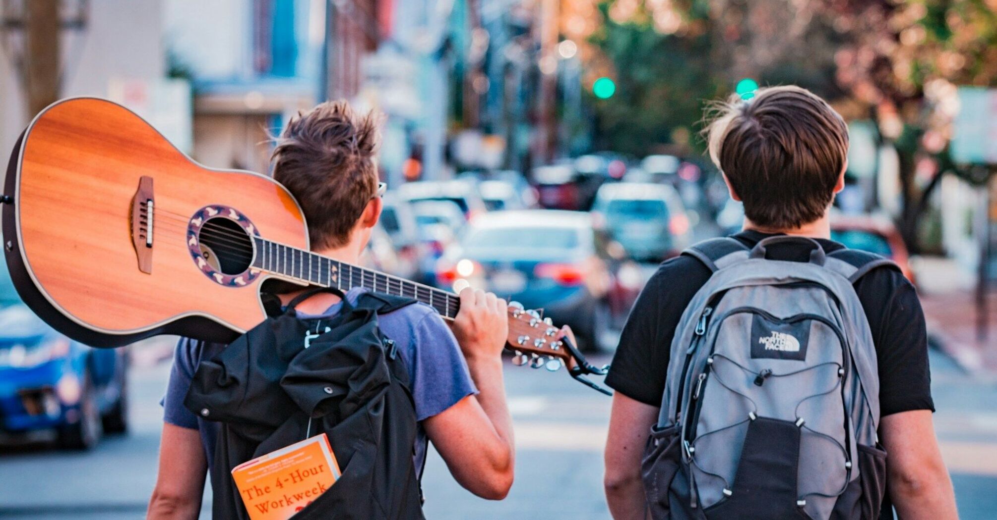 Two young travelers with backpacks, one carrying a guitar, walking down a street in a lively urban setting