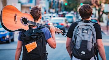 Two young travelers with backpacks, one carrying a guitar, walking down a street in a lively urban setting
