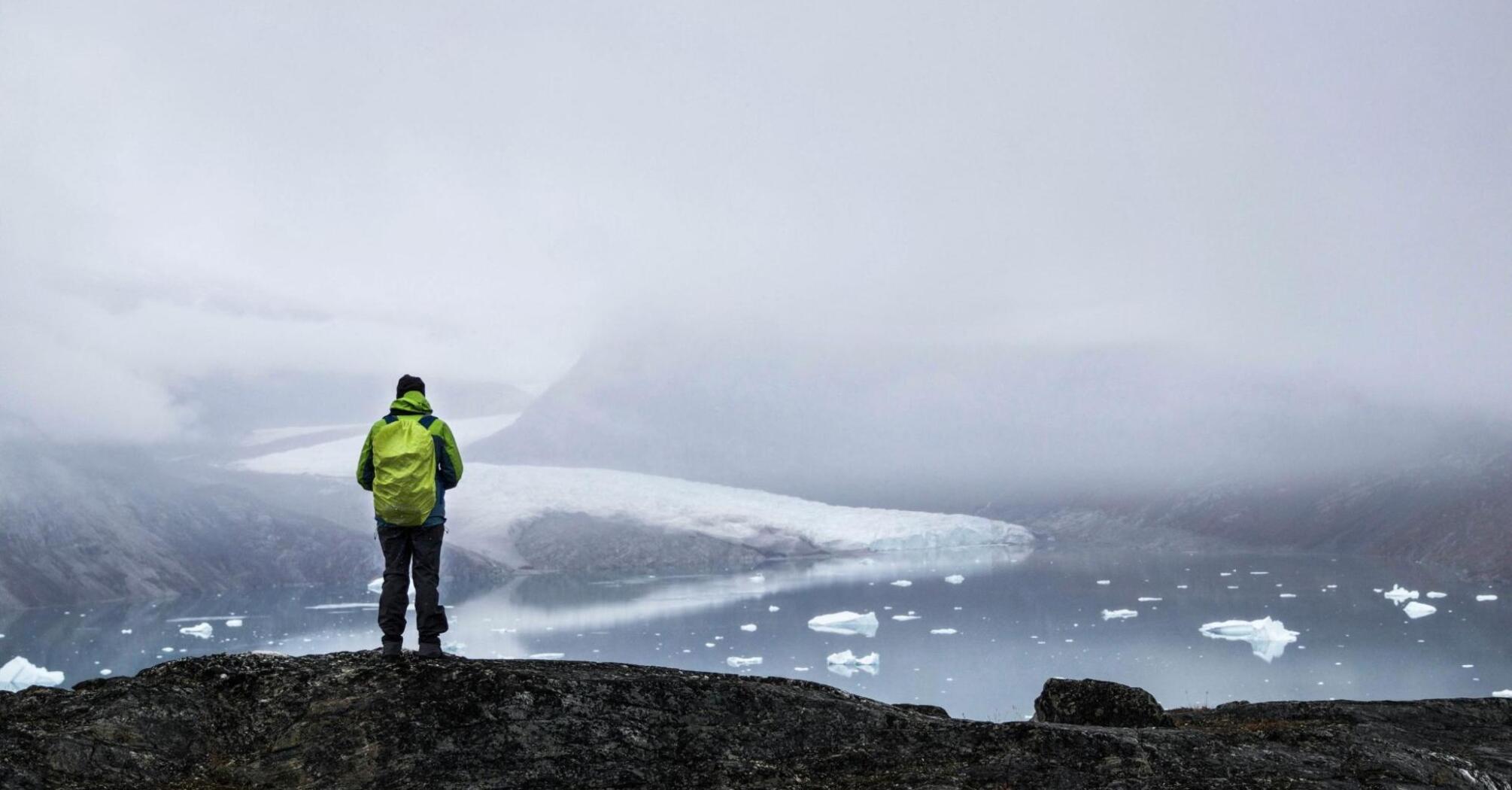 A hiker in a green jacket stands on a rocky outcrop, overlooking a misty Greenlandic landscape with icebergs and a glacier in the distance