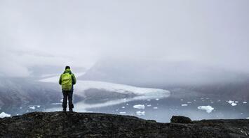 A hiker in a green jacket stands on a rocky outcrop, overlooking a misty Greenlandic landscape with icebergs and a glacier in the distance
