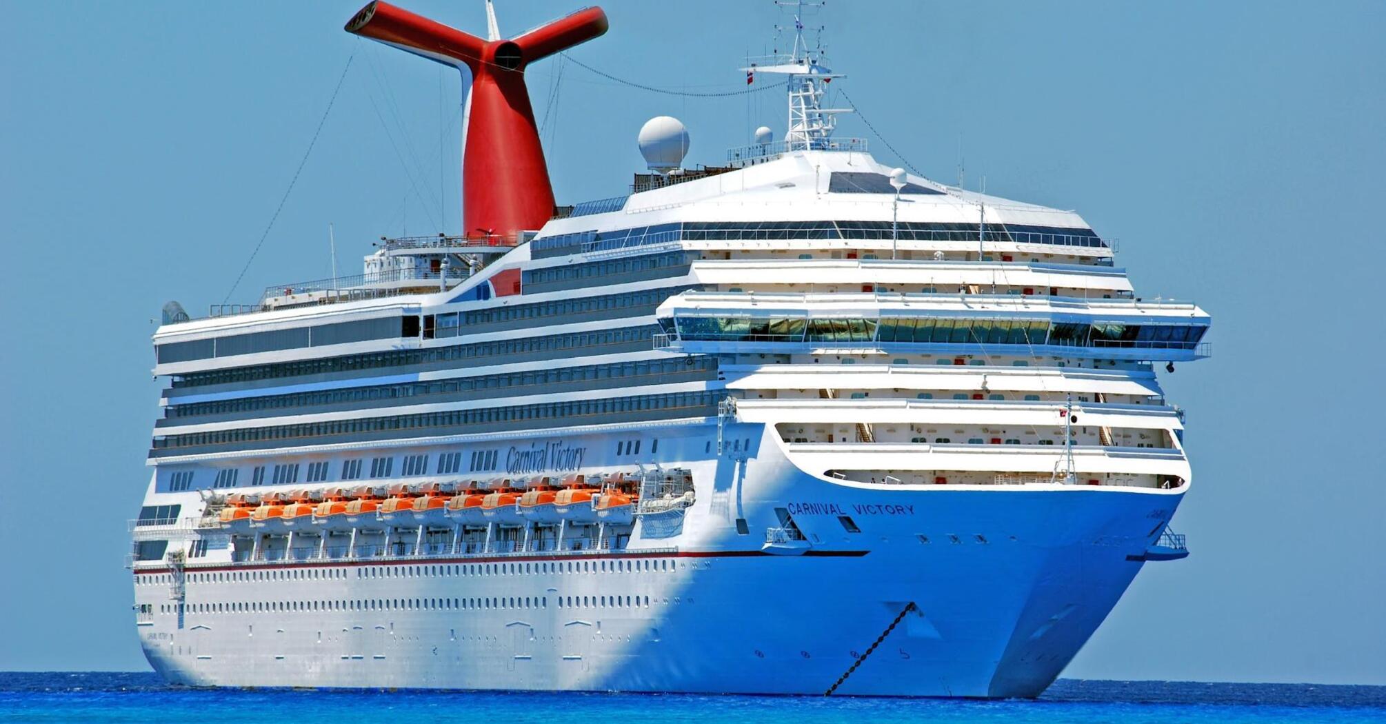 A large white cruise ship, Carnival Victory, sails through clear blue waters under a bright sky