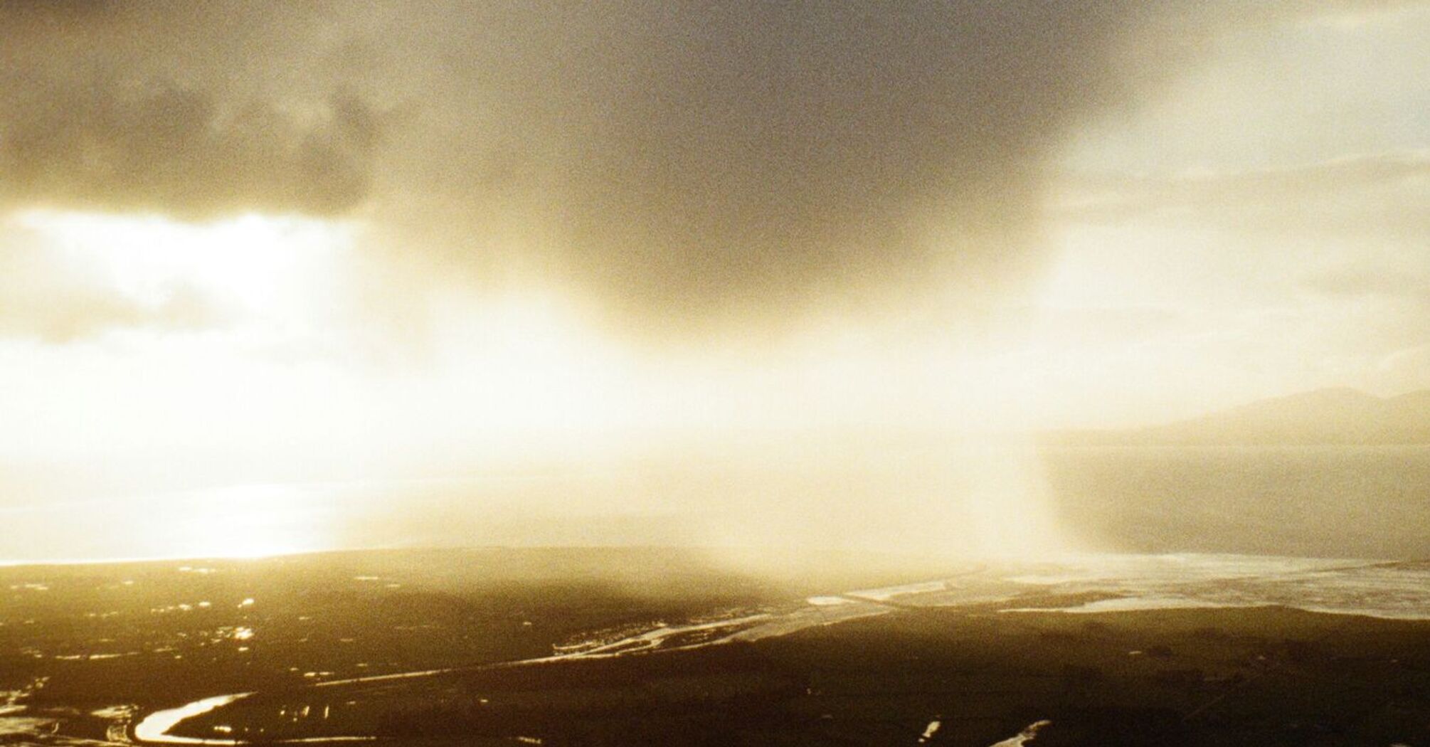 A dark storm cloud looms over a vast landscape with rivers winding through fields, reflecting the dim sunlight as the storm approaches