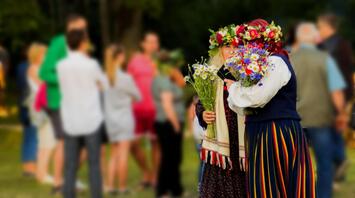 Two women in traditional clothing, adorned with floral wreaths, holding bouquets of wildflowers at an outdoor cultural event