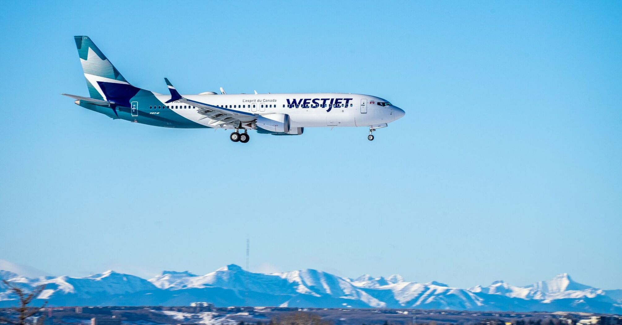 A WestJet airplane flying against a clear blue sky with snow-capped mountains in the background