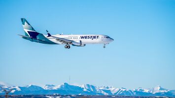 A WestJet airplane flying against a clear blue sky with snow-capped mountains in the background