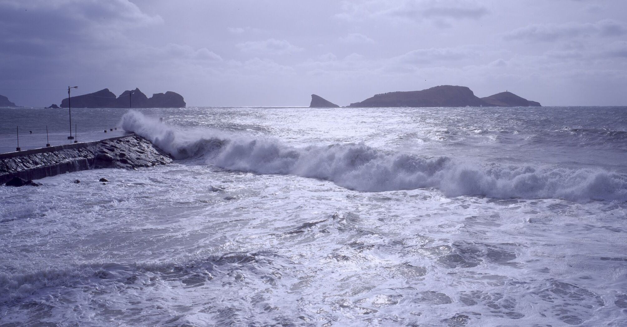 Waves crashing against a pier with distant islands in the background during a stormy sea