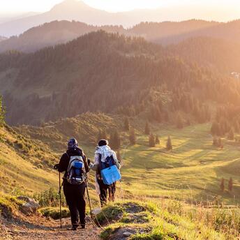Two hikers walking down a mountain trail at sunset with scenic views of rolling hills and pine trees