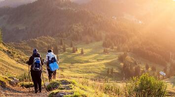 Two hikers walking down a mountain trail at sunset with scenic views of rolling hills and pine trees