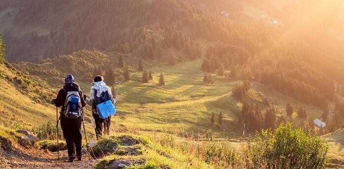 Two hikers walking down a mountain trail at sunset with scenic views of rolling hills and pine trees