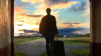 A silhouette of a person with a suitcase standing under an archway, gazing at a vibrant sunset over an open landscape