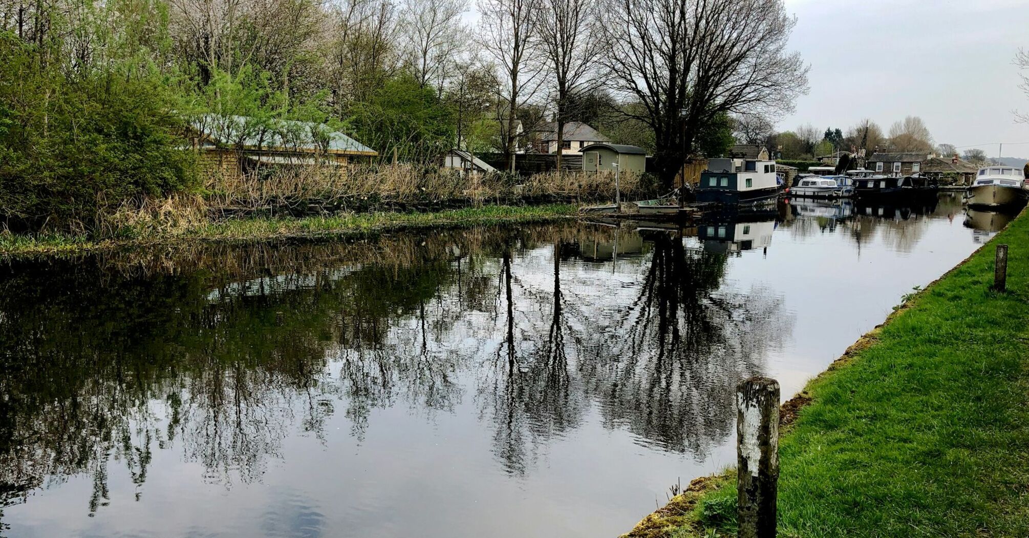 A calm canal with moored boats and trees reflected in the water, surrounded by greenery on a cloudy day