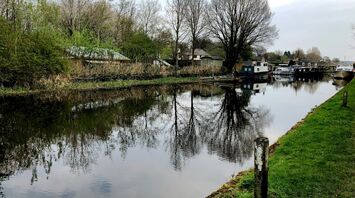 A calm canal with moored boats and trees reflected in the water, surrounded by greenery on a cloudy day