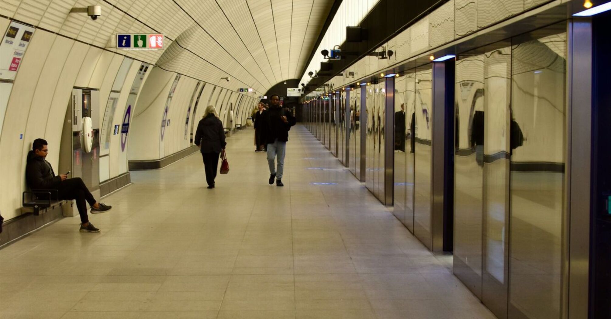 A quiet, spacious platform of the Elizabeth line with a few passengers walking and sitting, reflecting the modern design of the station