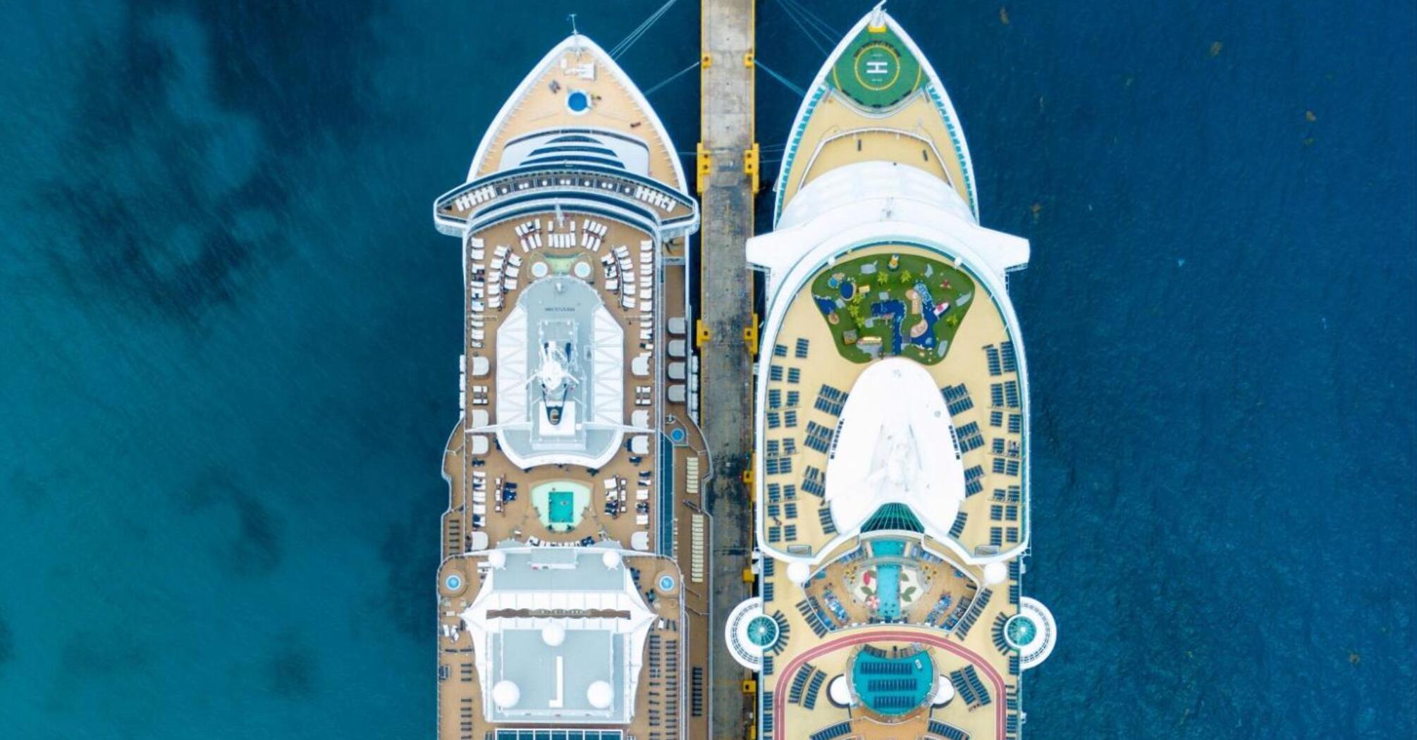 Aerial view of two cruise ships docked side by side at a pier, showcasing their large decks with pools and amenities, surrounded by deep blue ocean water