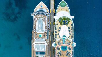 Aerial view of two cruise ships docked side by side at a pier, showcasing their large decks with pools and amenities, surrounded by deep blue ocean water