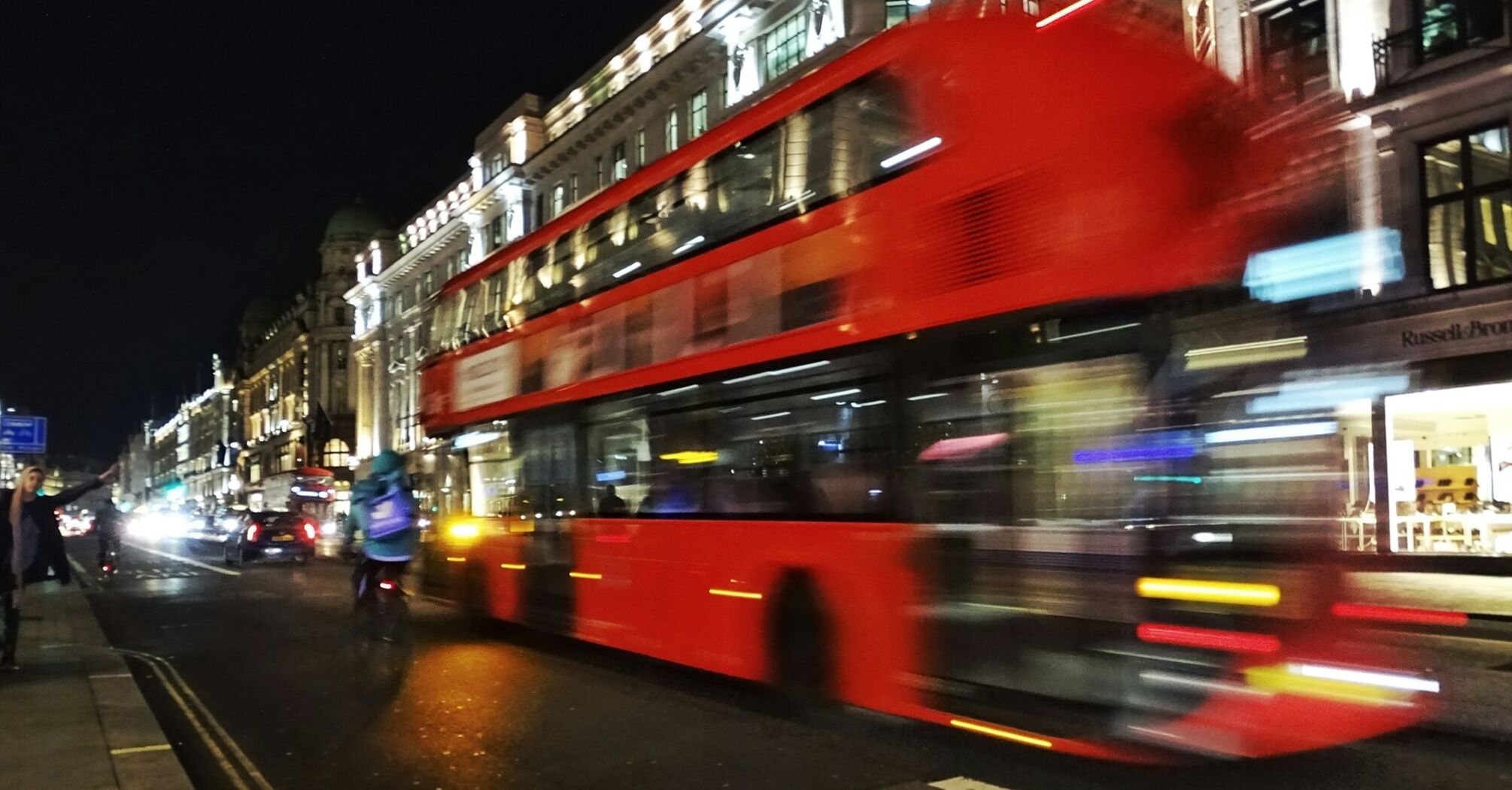 Blurred image of a red London double-decker bus speeding down a busy, illuminated street at night