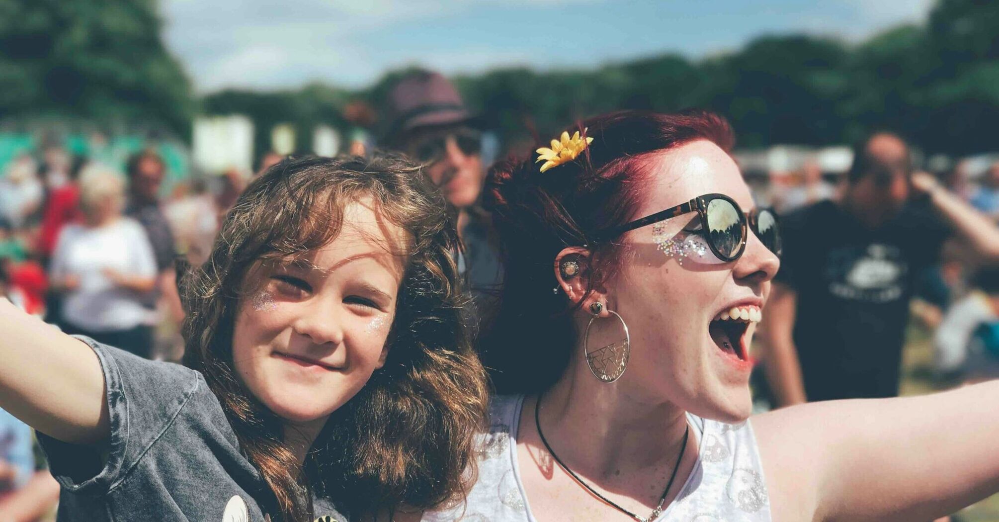 A joyful woman with sunglasses and a flower in her hair smiles widely while hugging a young girl with curly hair at an outdoor event