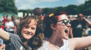A joyful woman with sunglasses and a flower in her hair smiles widely while hugging a young girl with curly hair at an outdoor event