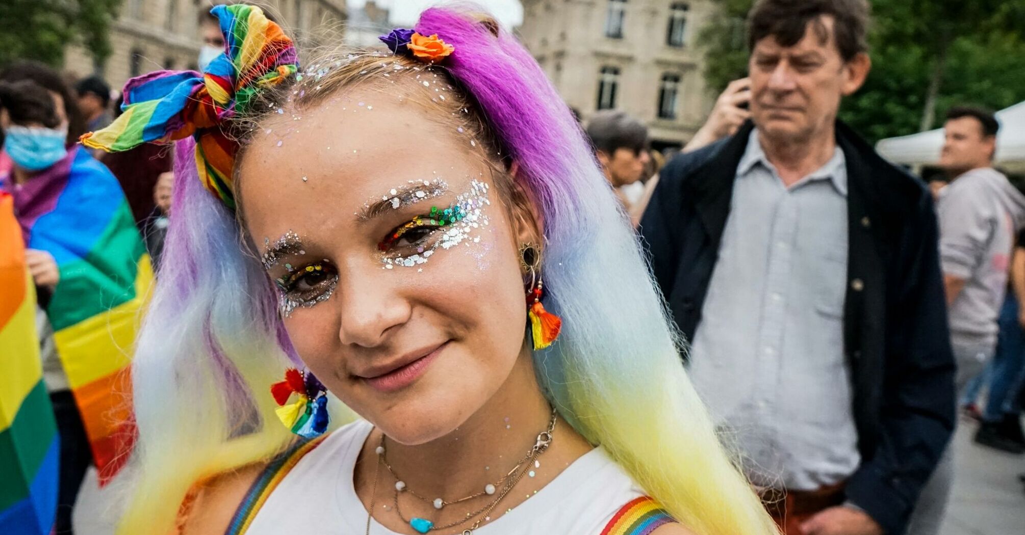 A young person with rainbow-colored hair and glitter makeup smiling at a Pride event