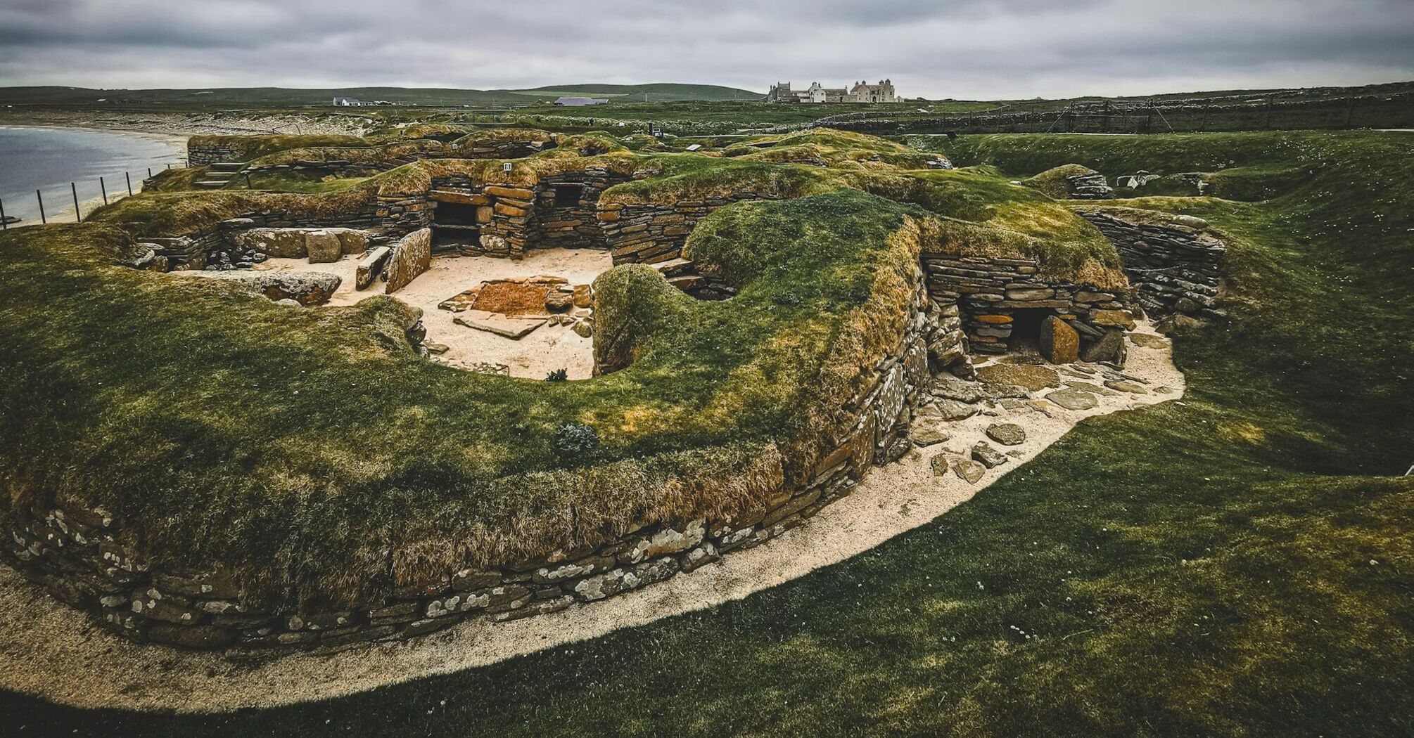 Ancient stone structures of Skara Brae in Orkney, with a cloudy sky and coastline in the background