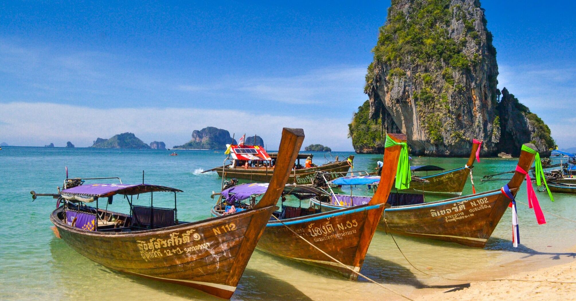 Traditional Thai longtail boats anchored by a scenic limestone cliff on a sunny day