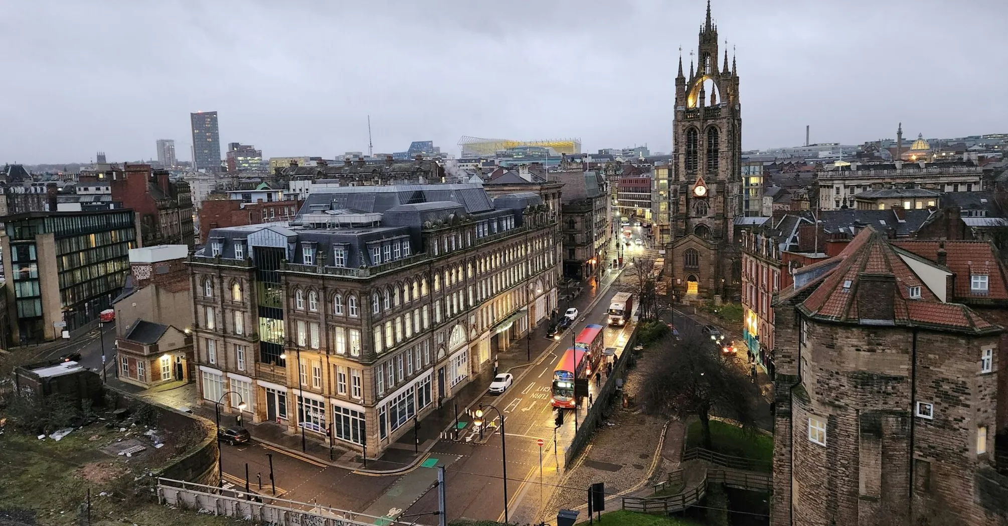 A view of a Newcastle city street in the early evening with historic buildings, buses, and a prominent church tower in the background, under a cloudy sky